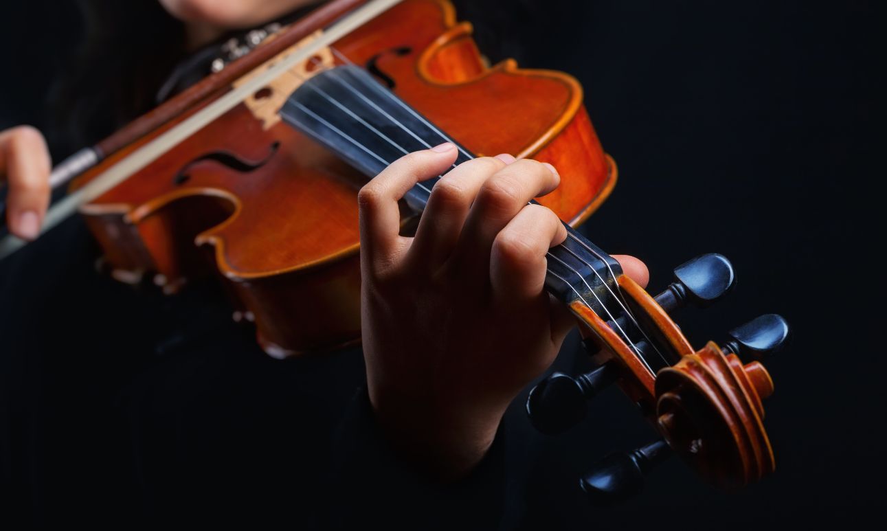 Close-up of hands playing a violin against a dark background. The musician's fingers press the strings, conveying a sense of focus and musical artistry.
