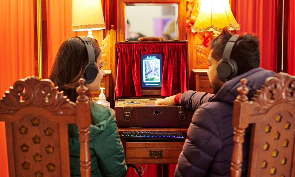 Two people facing away, sitting on wooden chairs, watch a small vintage TV. The room is warmly lit with a nostalgic atmosphere, featuring red curtains and lamps.