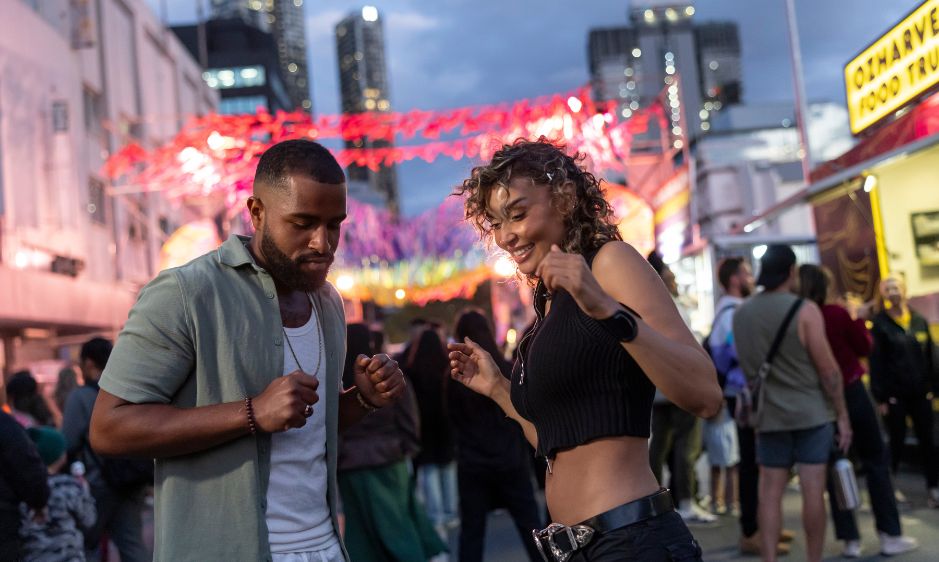 A cheerful pair poses in front of vibrant neon lights at a lively carnival. The scene is bustling with people, creating an energetic and festive atmosphere.