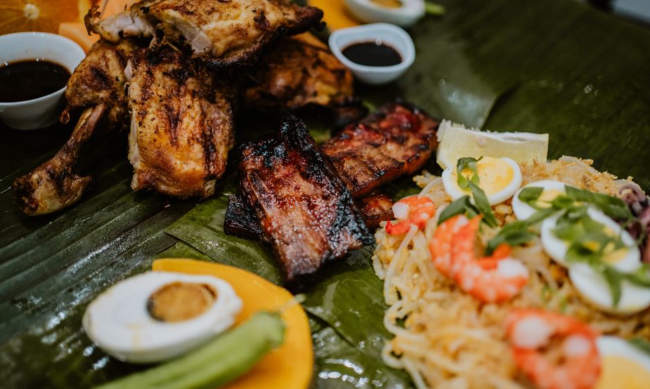 Close-up of a vibrant Filipino feast on banana leaves, featuring grilled chicken, ribs, pancit noodles with shrimp and eggs, and dipping sauces.