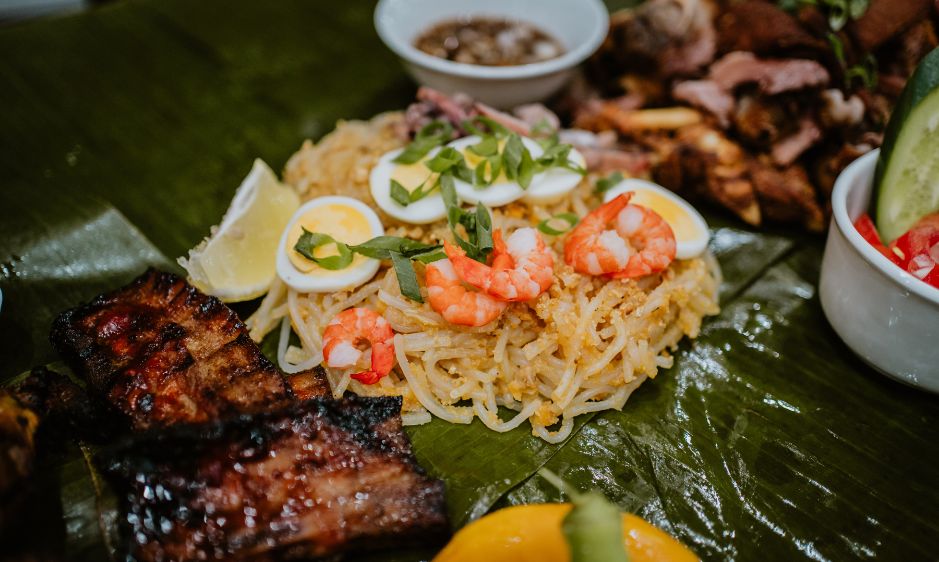 Close-up of a vibrant Filipino feast on banana leaves, featuring grilled chicken, ribs, pancit noodles with shrimp and eggs, and dipping sauces.