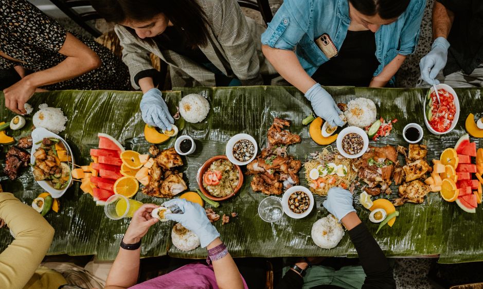 Top view of people wearing gloves sharing a vibrant meal on a banana leaf-covered table, featuring grilled meats, fruits, and various sauces. Lively and communal.