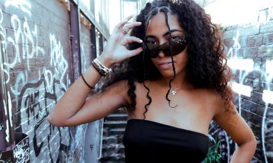 Young woman with curly hair in a black strapless top, playfully adjusting sunglasses. She's standing against a graffiti-covered wall, exuding a confident vibe.
