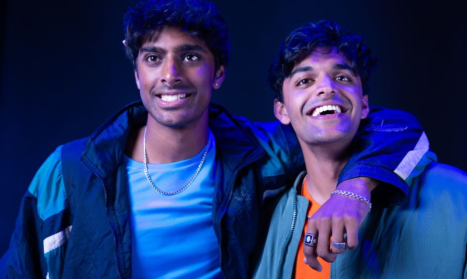 Two friends stand closely, smiling under blue lighting. One has an arm around the other. They wear casual jackets, creating a warm, friendly atmosphere.