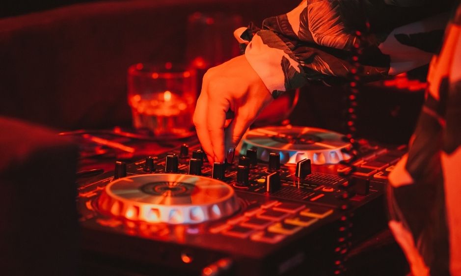 A DJ adjusts a mixer in a dimly lit club, bathed in warm red light. A hand delicately turns dials, with a glass in the background, creating an energetic atmosphere.