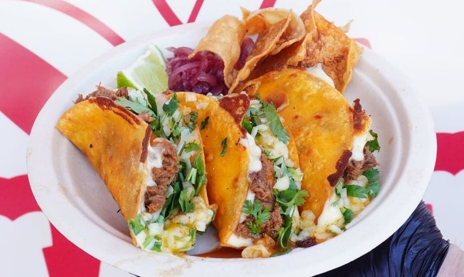 A plate of crispy tacos filled with meat, lettuce, and cheese is accompanied by tortilla chips and salsa. The setting is bright, creating a fresh and appetizing feel.