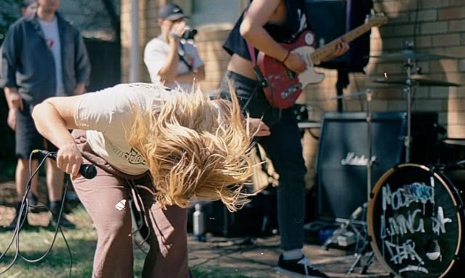 A woman energetically headbanging at an outdoor concert with a guitarist in the background and festival equipment nearby, conveying excitement and vibrancy.