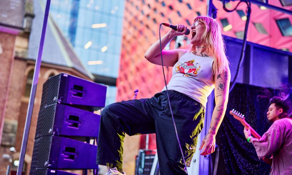 Singer performs energetically on stage, wearing a white tank top and jeans, with colorful lights and a supporting guitarist in the background, creating a lively atmosphere.
