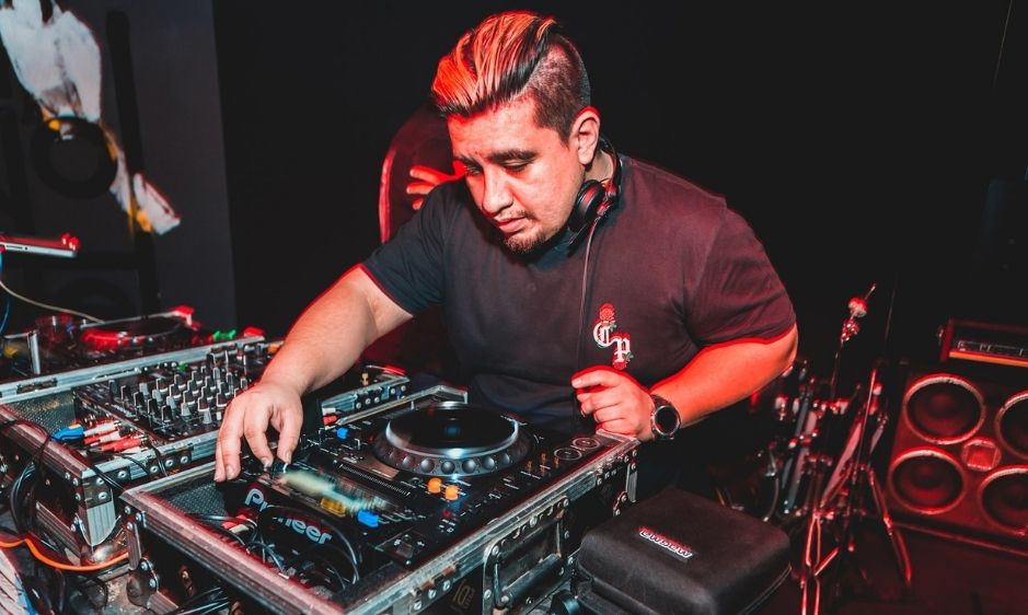 A DJ wearing a black shirt focuses intently on mixing music at a soundboard. The scene is dimly lit with red lighting, creating an energetic atmosphere.