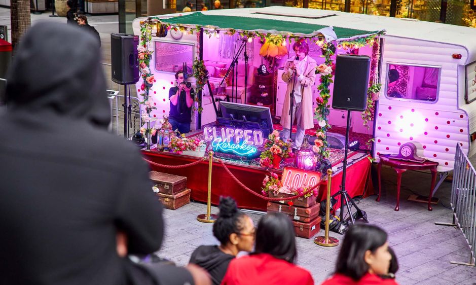 A person in a long coat performs on a brightly lit stage with pink and purple hues. A neon sign reads "CLIPPED," surrounded by floral decorations.