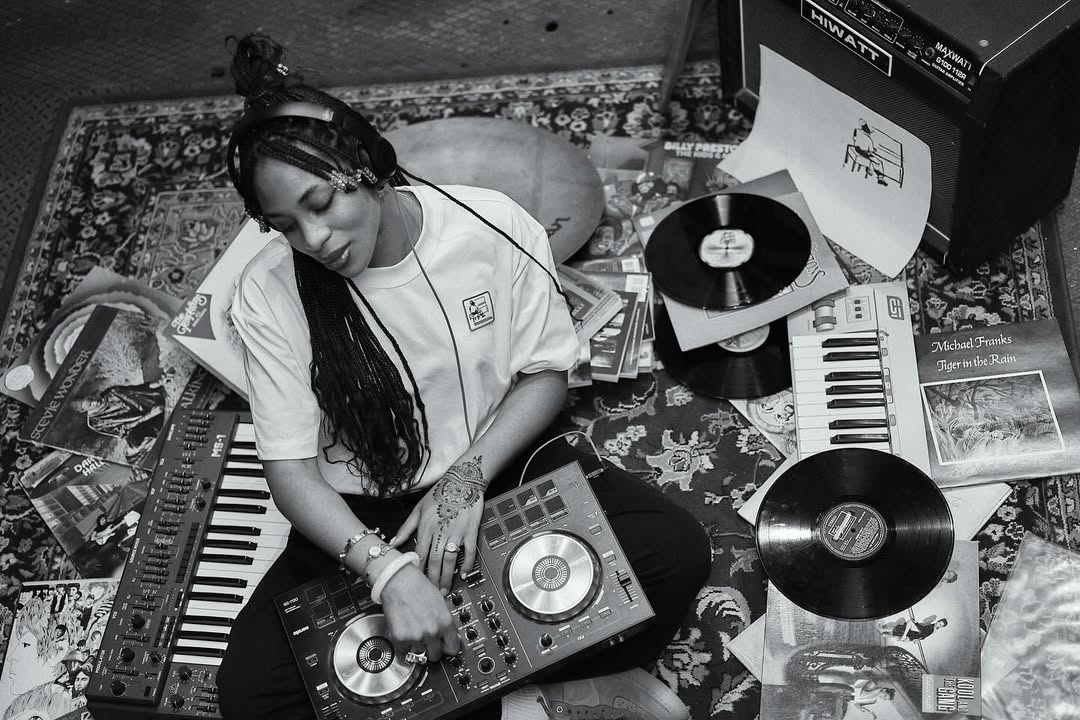 Black and white image of a woman sitting on a rug, surrounded by vinyl records and a turntable. She wears headphones, smiling, creating a nostalgic, relaxed vibe.