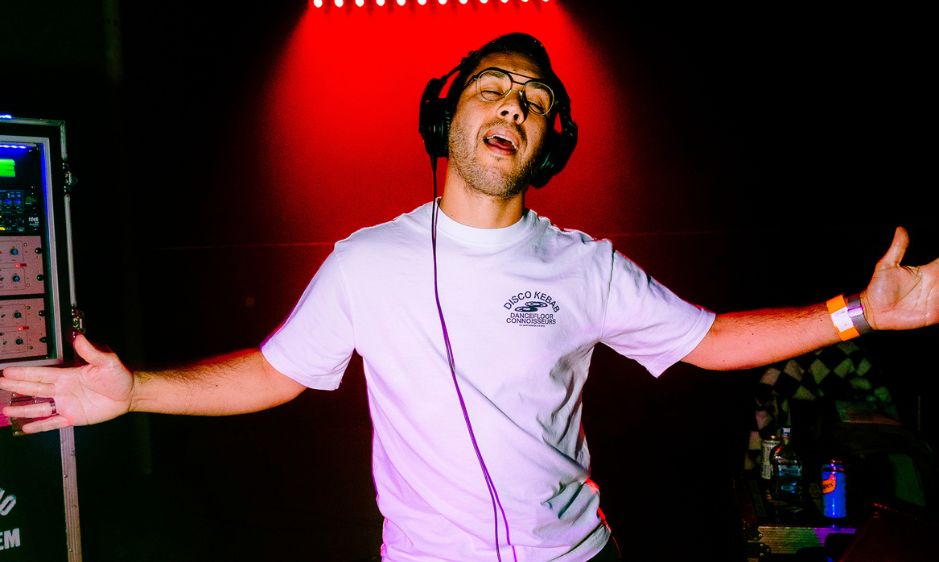 A person with curly hair joyfully spreads their arms wide under a bright red spotlight. They wear a casual white T-shirt, exuding excitement and energy.