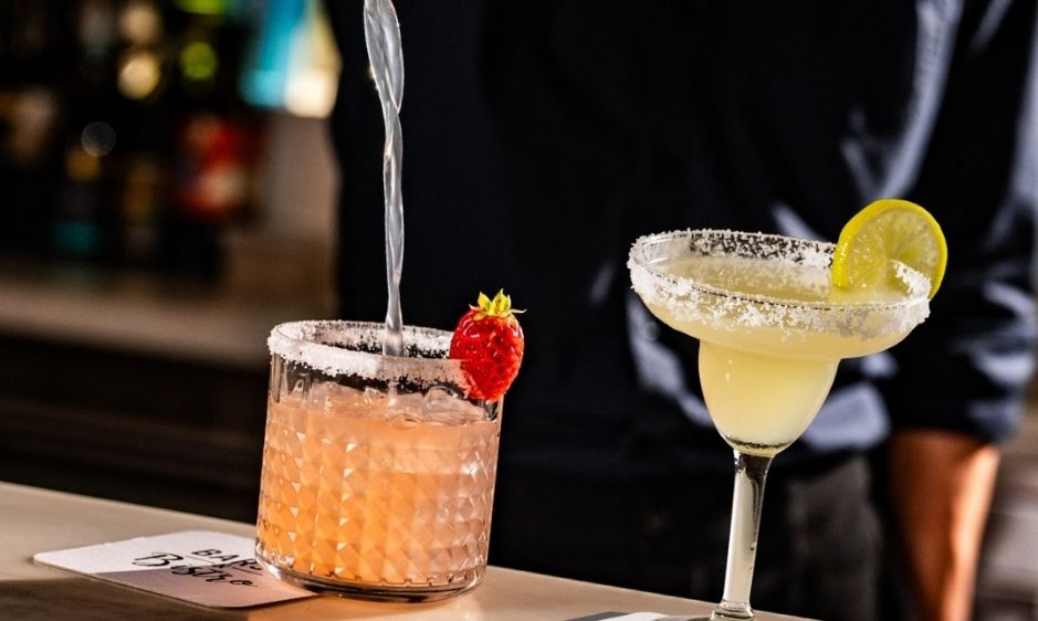 Two cocktails on a bar: a pink drink with a strawberry and a margarita with a lime slice. A bartender stands behind, creating a relaxed atmosphere.