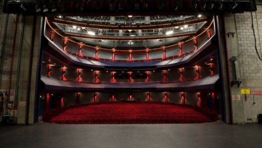 View of an ornate theater from the stage, showcasing a red carpeted floor, three levels of seating, and gilded architectural details, exuding elegance.
