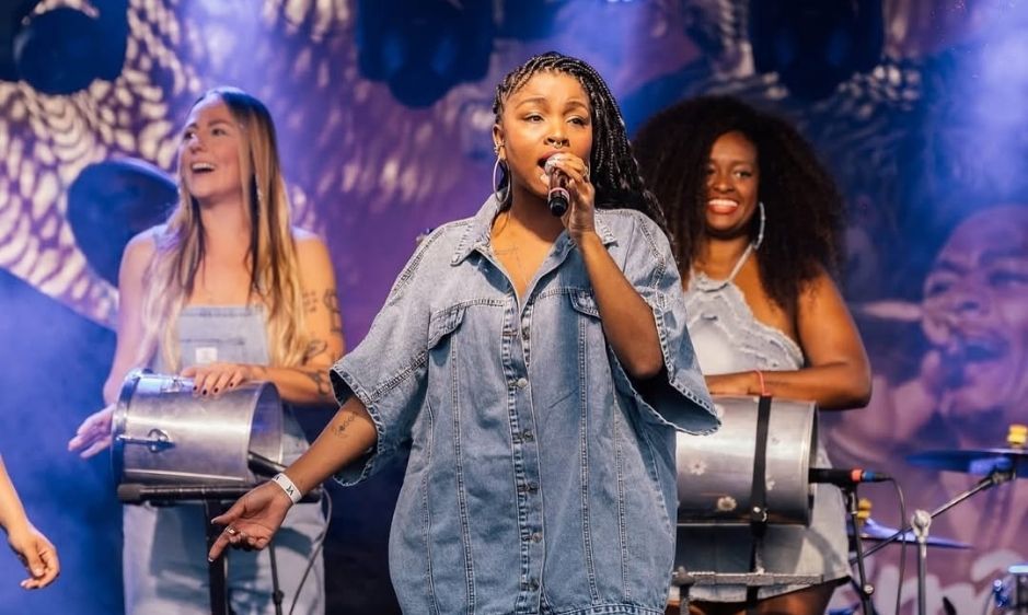 Three performers on stage, with the central person singing into a microphone wearing denim. Two others play percussion instruments, smiling and energetic.