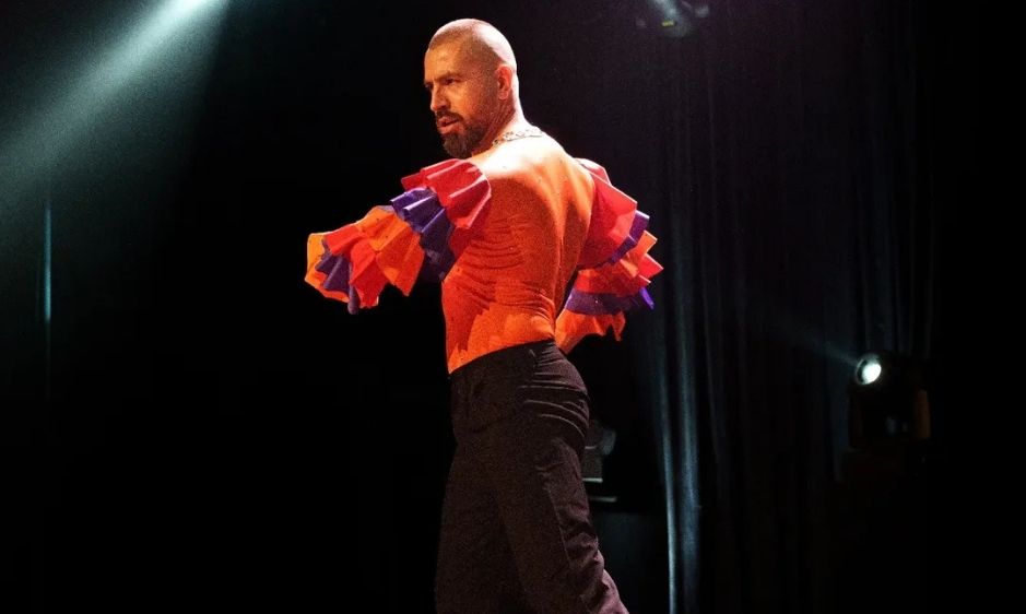 A dancer in a red and black costume strikes a pose on stage under bright spotlight beams, conveying passion and intensity in a dark theater setting.