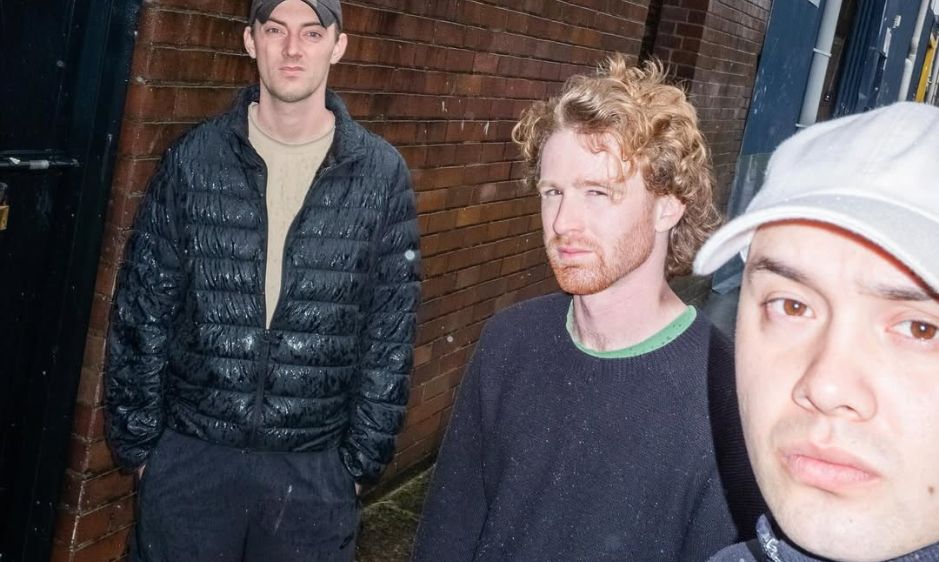 Three people are standing in an alley at night. One wears a black jacket, another a sweater, and the third a cap. The mood seems casual and candid.