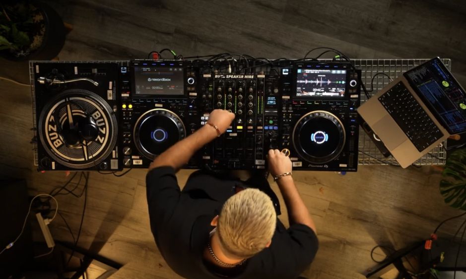 Overhead view of a DJ with shaved head mixing music on a turntable setup. The dim lighting and focused stance convey concentration and rhythm.