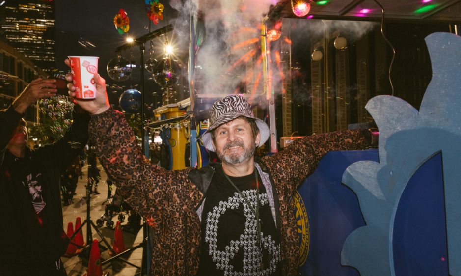 A man wearing a leopard print coat and patterned hat holds up a drink cup, smiling. He stands in a colorful street scene at night with festive lights and decorations, conveying a joyful atmosphere.