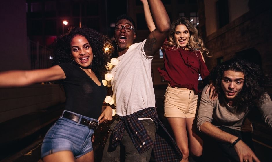 A group of four friends joyfully pose in a dimly lit urban street at night. They smile and laugh, capturing a lively, carefree atmosphere.