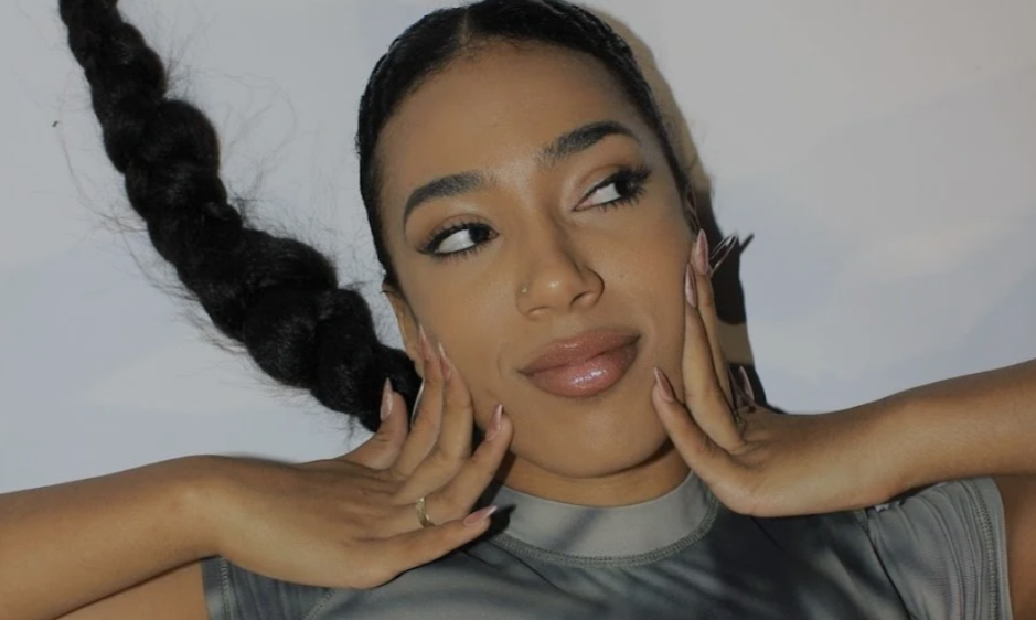 A woman with a long braided ponytail lies on a white surface, wearing a grey shirt. She has a serene expression with her hands gently on her cheeks.