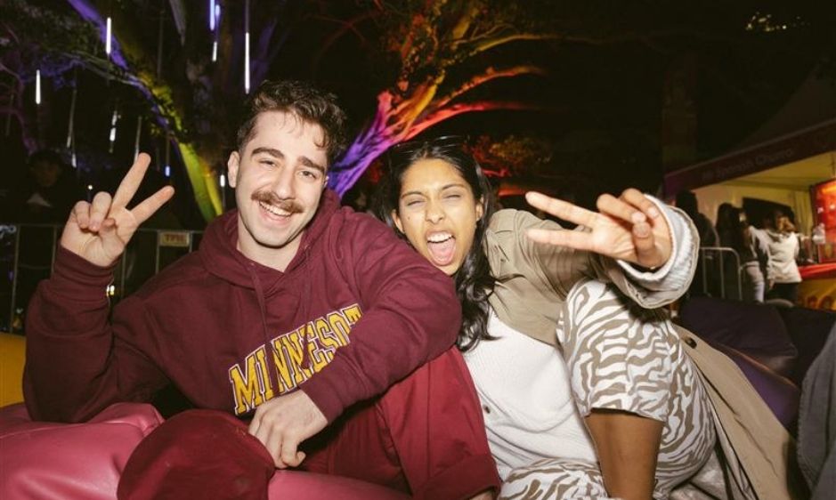 A smiling group of people hold up peace signs. They are seated outdoors at night with colourful lights and trees in the background, creating a festive vibe.