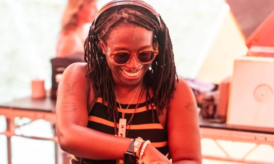 A person with braided hair and sunglasses dances joyfully in a bright room. They wear a striped top and bracelets, conveying a fun, lively atmosphere.