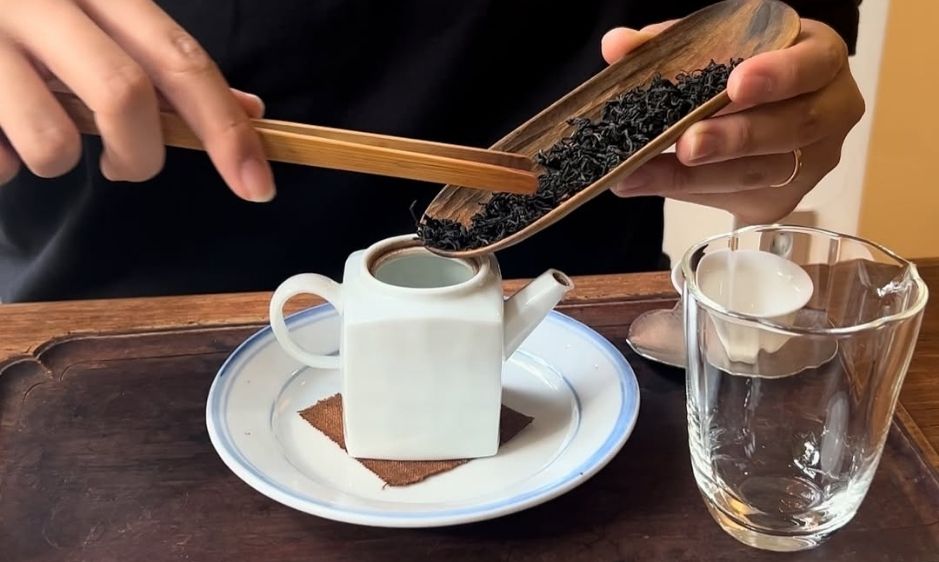 A hand grates chocolate over a cup of coffee on a white saucer. A glass of water is nearby, creating a cozy and inviting atmosphere on a wooden table.