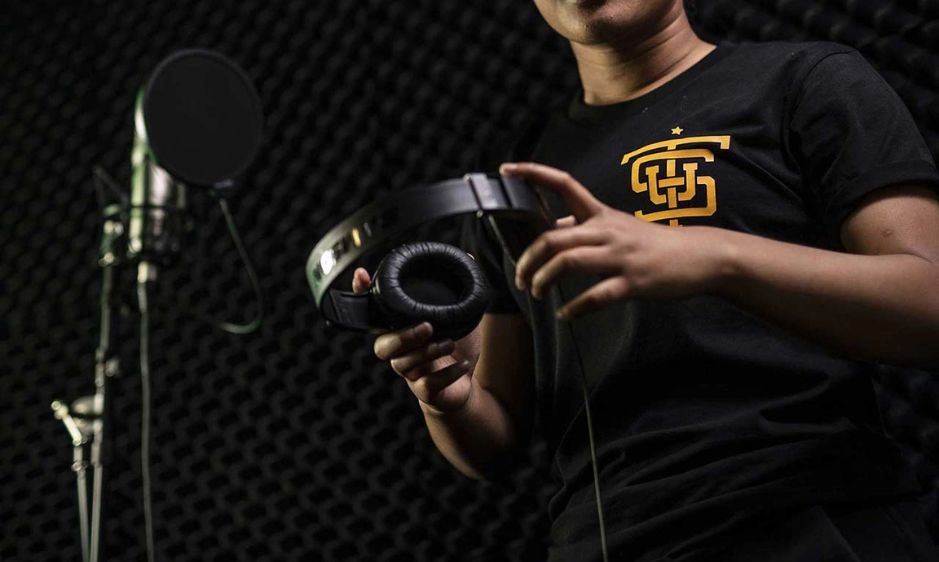 A person in a dark recording studio holds headphones near a microphone. The atmosphere is focused and creative, suggesting music production.