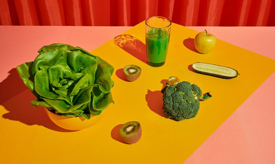 Assorted vegetables and fruits, including lettuce, kiwi, broccoli, cucumber, lemon, and bell pepper, rest on an orange surface with a red curtain backdrop. The scene is colorful and vibrant.