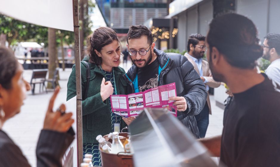 A group of people enjoy donuts from a street vendor. Two people hold pink donut boxes while smiling, creating a joyful and lively atmosphere.