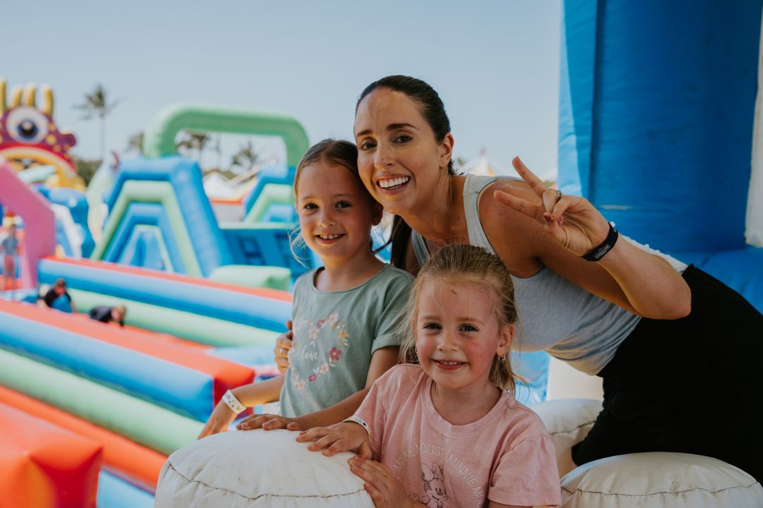 A woman and two children smile and pose at a colorful inflatable playground. The scene is bright and cheerful, conveying a sense of fun and joy.