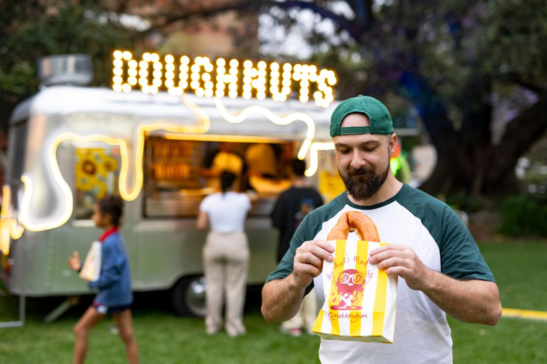 Man in a green cap smiles while holding a bag of doughnuts. A food truck labeled "Doughnuts" and customers are visible in the background at a park.
