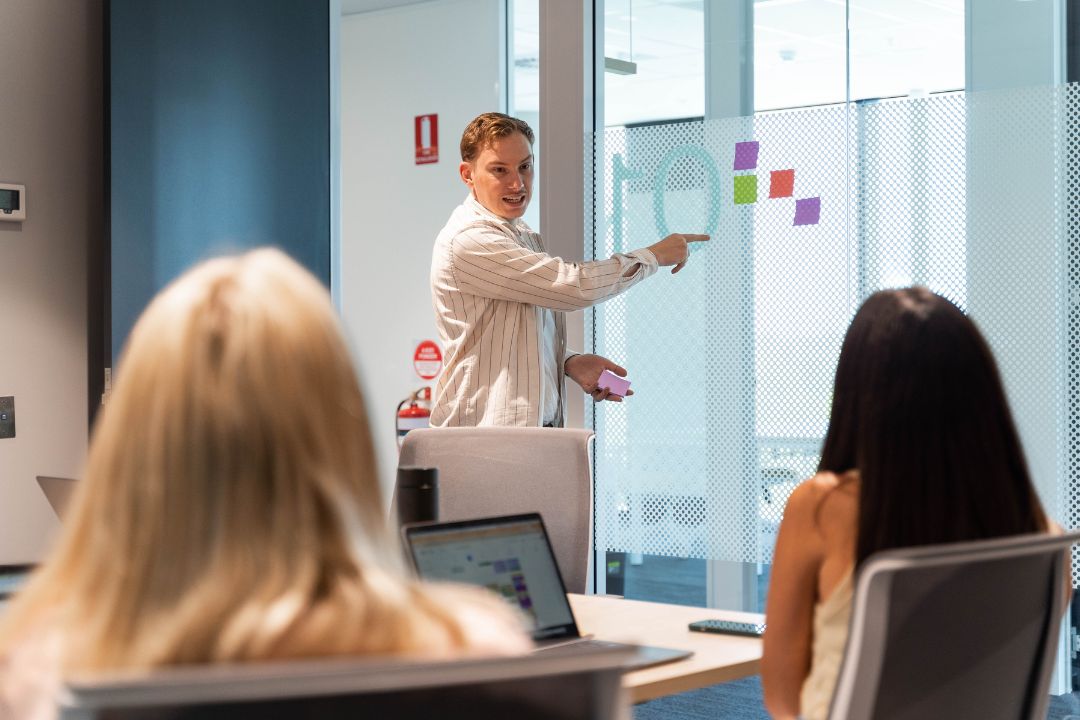 A person in a business setting presents at a whiteboard with colorful charts, engaging two seated colleagues. The atmosphere is focused and professional.