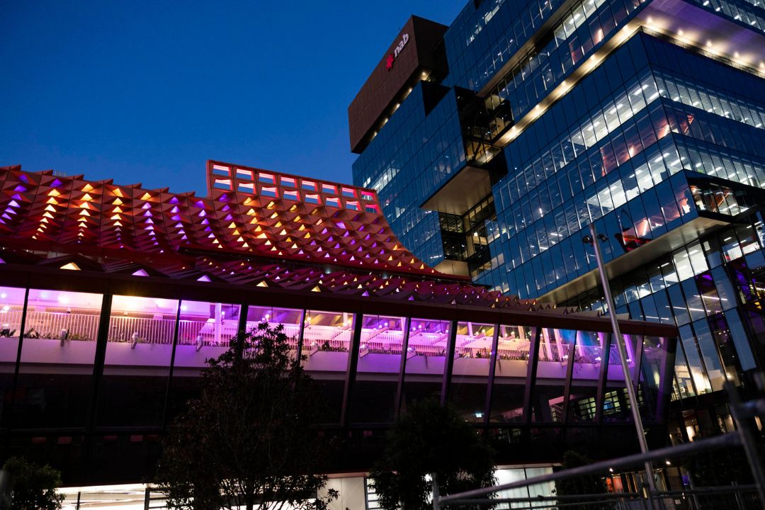 Modern building with vibrant purple and red lights against a deep blue sky. The atmosphere is lively and urban, with reflections on glass facades.