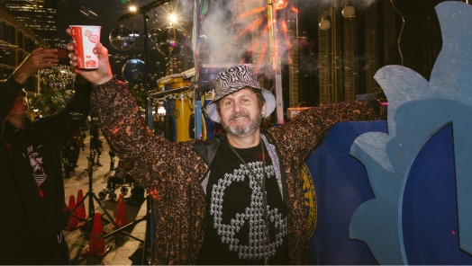 A man wearing a leopard print coat and patterned hat holds up a drink cup, smiling. He stands in a colorful street scene at night with festive lights and decorations, conveying a joyful atmosphere.