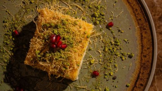 A square piece of kunafa on a green plate, topped with crushed pistachios and pomegranate seeds. The plate has scattered pistachios and seeds.