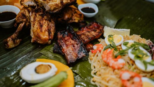 Close-up of a vibrant Filipino feast on banana leaves, featuring grilled chicken, ribs, pancit noodles with shrimp and eggs, and dipping sauces.