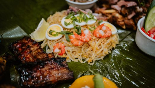 Close-up of a vibrant Filipino feast on banana leaves, featuring grilled chicken, ribs, pancit noodles with shrimp and eggs, and dipping sauces.
