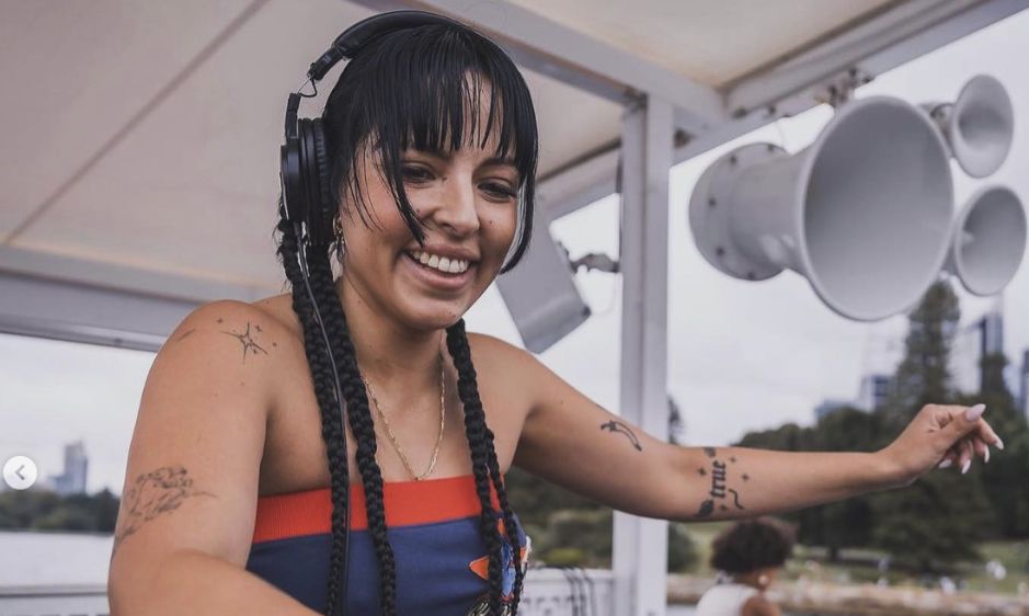 A smiling DJ with braided hair and tattoos, wearing headphones, mixes music on a deck at an outdoor event.