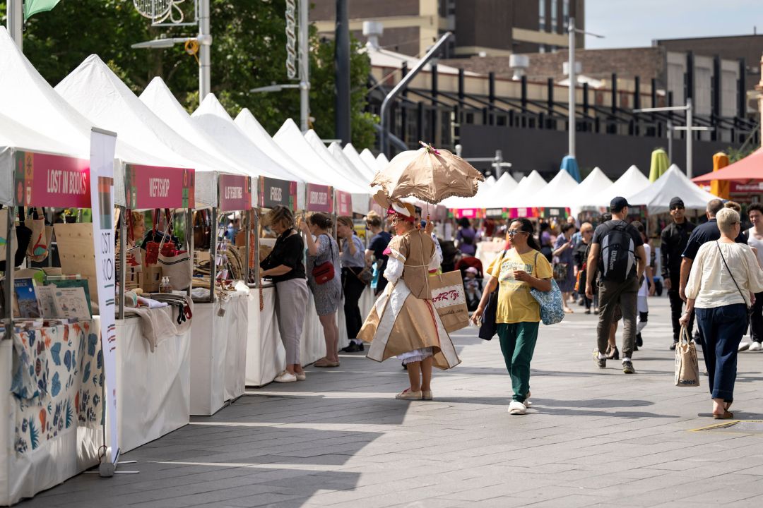 A bustling outdoor market with white tents showcasing art and crafts. A person in a floral costume walks among casually dressed people. Lively and festive.