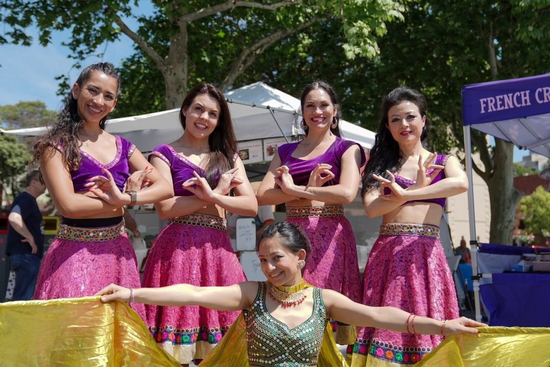 Five young performers in colourful costumes pose joyfully outdoors, under a sunny sky. Lush trees and event tents are visible in the background.