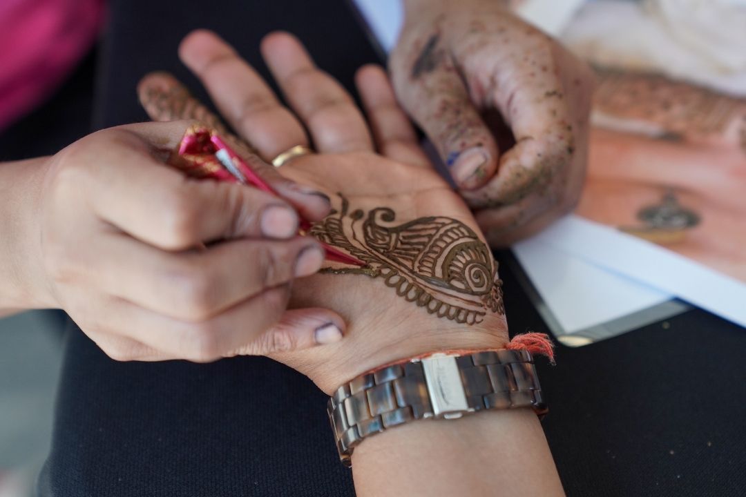 A person applies intricate henna designs on another's palm, focusing on a detailed floral pattern; the mood is artistic and focused.