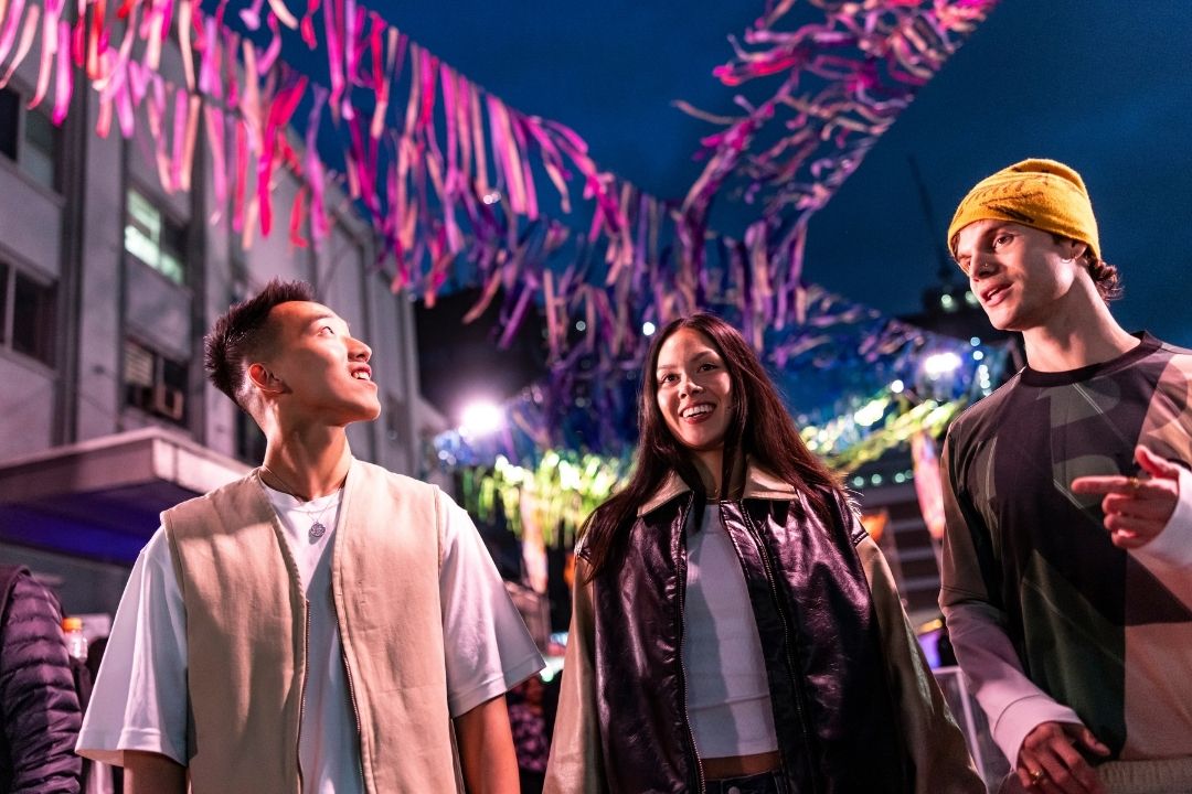 Three people enjoy a festive street scene, surrounded by colorful hanging ribbons and vibrant lights. The atmosphere is joyful and lively.