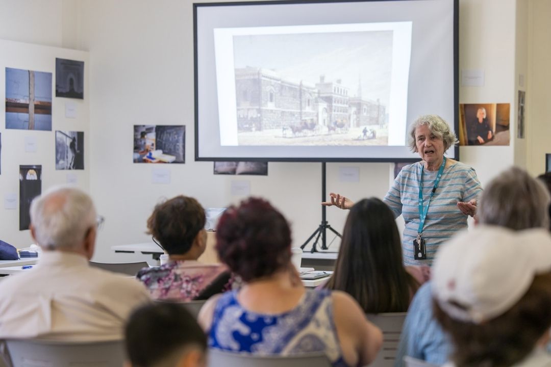 A lecturer presents to an engaged audience in a small room, featuring historical photos on the walls and a projected image. The atmosphere is informative and attentive.