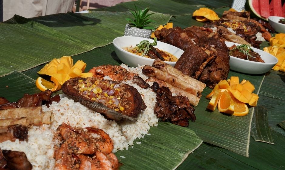 A traditional Asian meal served on banana leaves features rice, grilled meats, vegetables, and sliced mango, creating a vibrant and festive table setting.