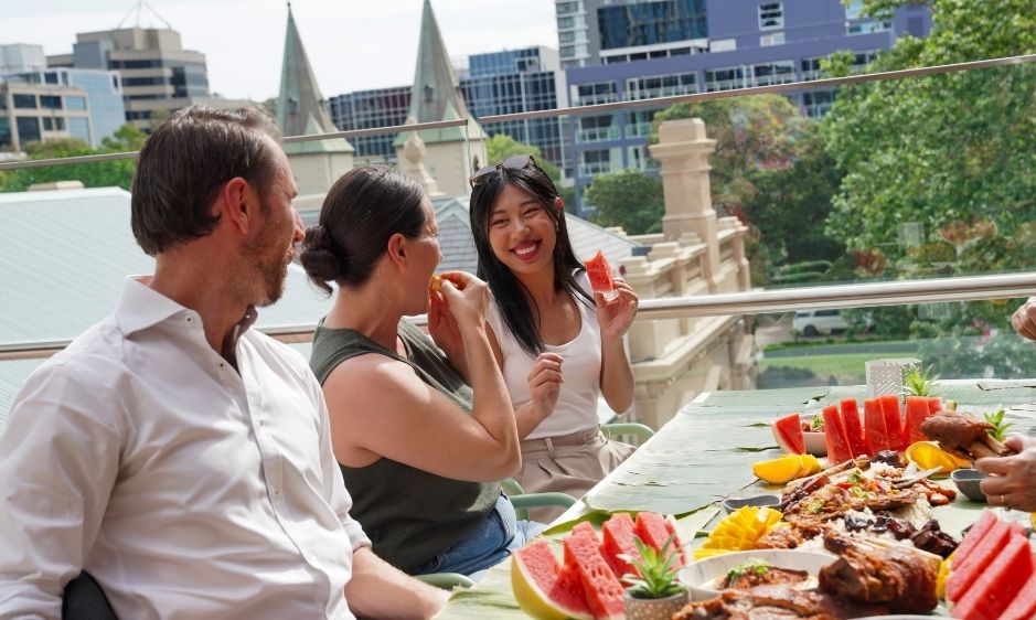 Three people enjoying a cheerful outdoor gathering with a table full of colourful fruit and snacks. A cityscape and greenery are visible in the background.