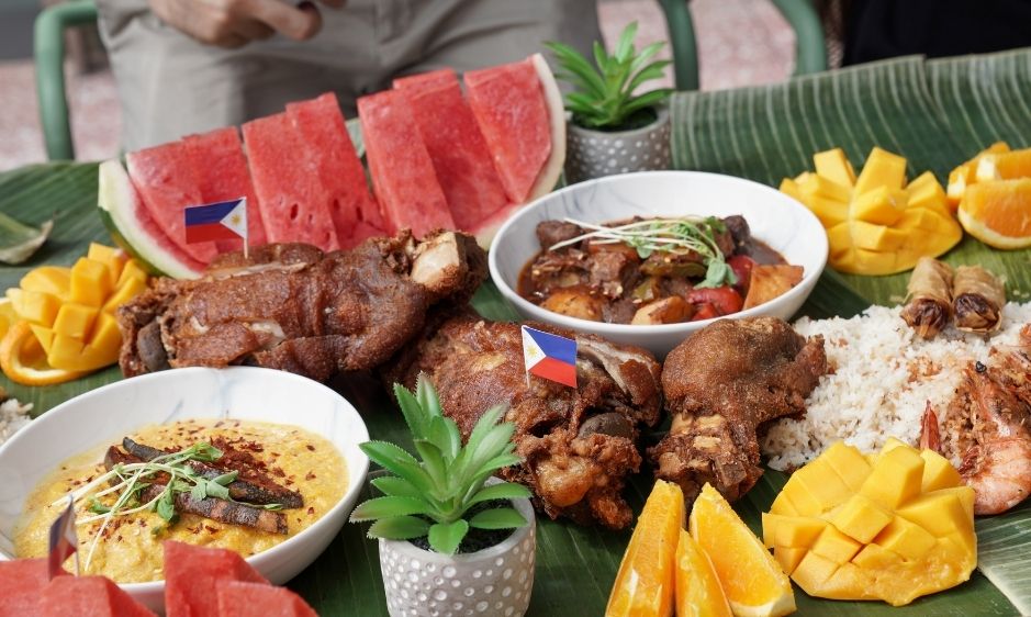 A vibrant feast on a banana leaf includes sliced watermelon, grilled chicken, mixed stir-fry, rice, and mango. Tropical and inviting atmosphere.