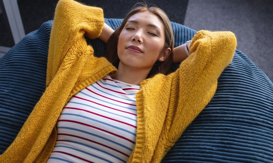 A woman relaxes peacefully on a dark blue bean bag chair, wearing a yellow cardigan and striped shirt, suggesting comfort and tranquillity.