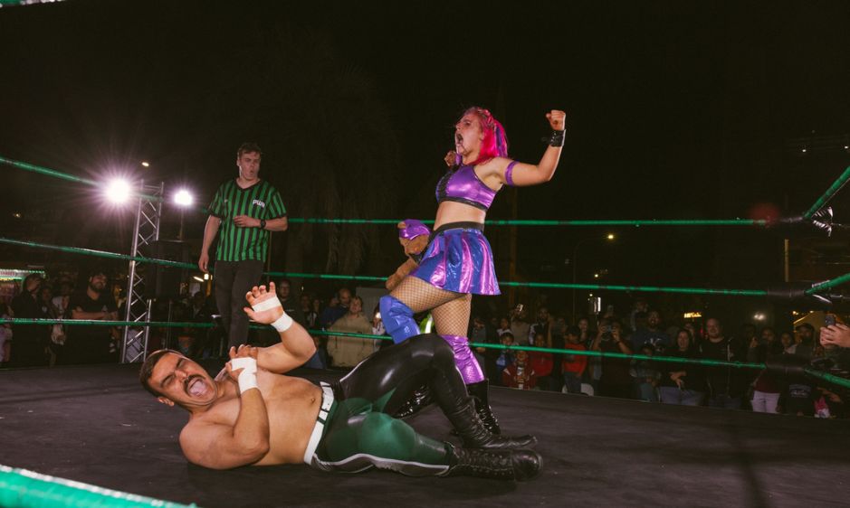 A wrestler in a purple outfit triumphantly poses in a ring, while her opponent, dressed in brown, lies on the mat. The scene is dynamic and energetic.